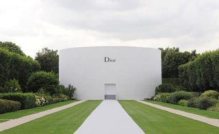 Outside, daytime image, cut lawn, stone pathways leading to a whiye curved Dior building, shrubs hedges, trees to the right and left, grey cloudy sky