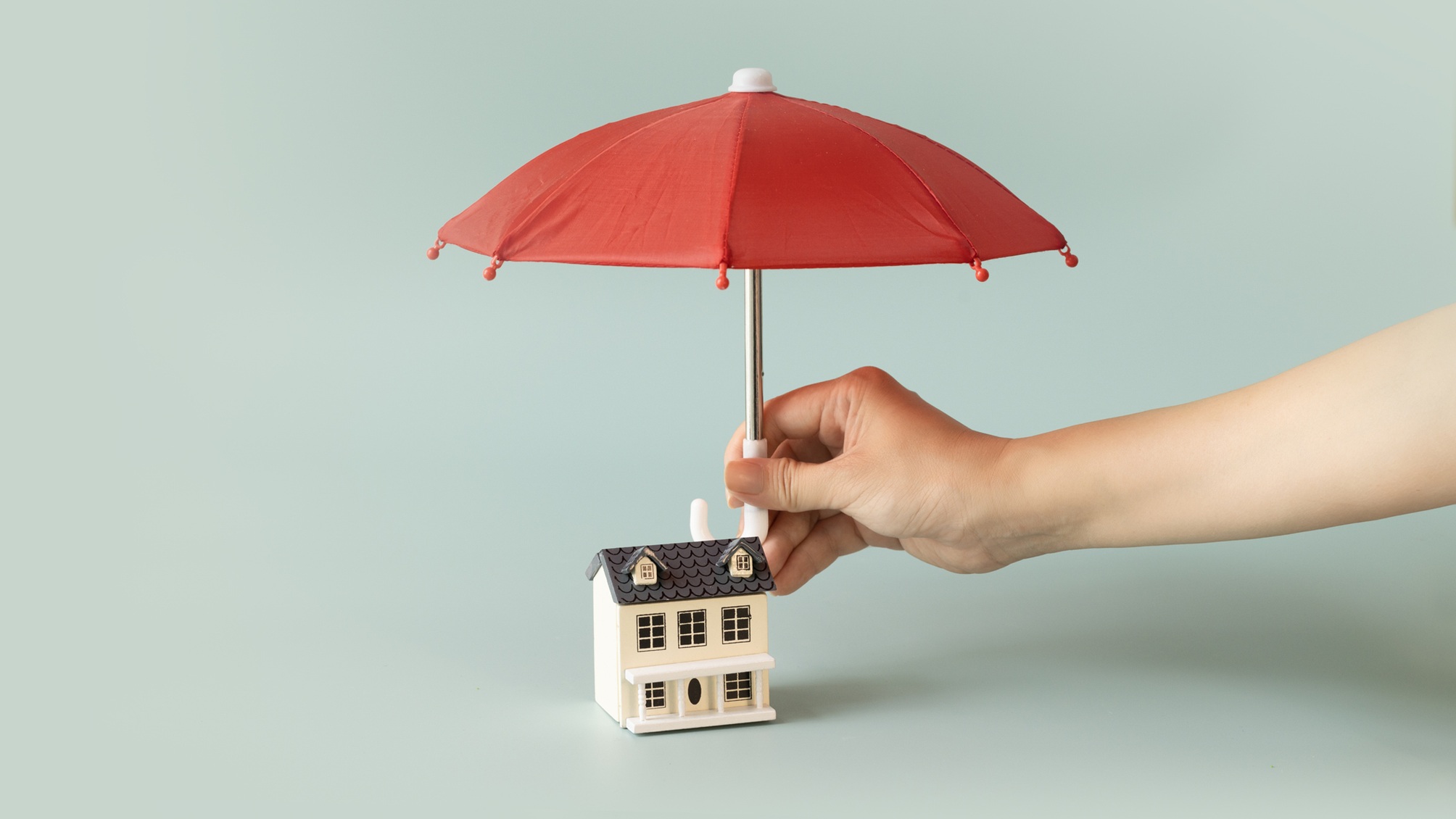 A hand holds a red umbrella over a toy home.