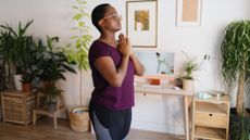 Woman stands in living room with her hands in prayer position and eyes closed