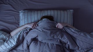 A person lies in bed with their covers pulled up over their head