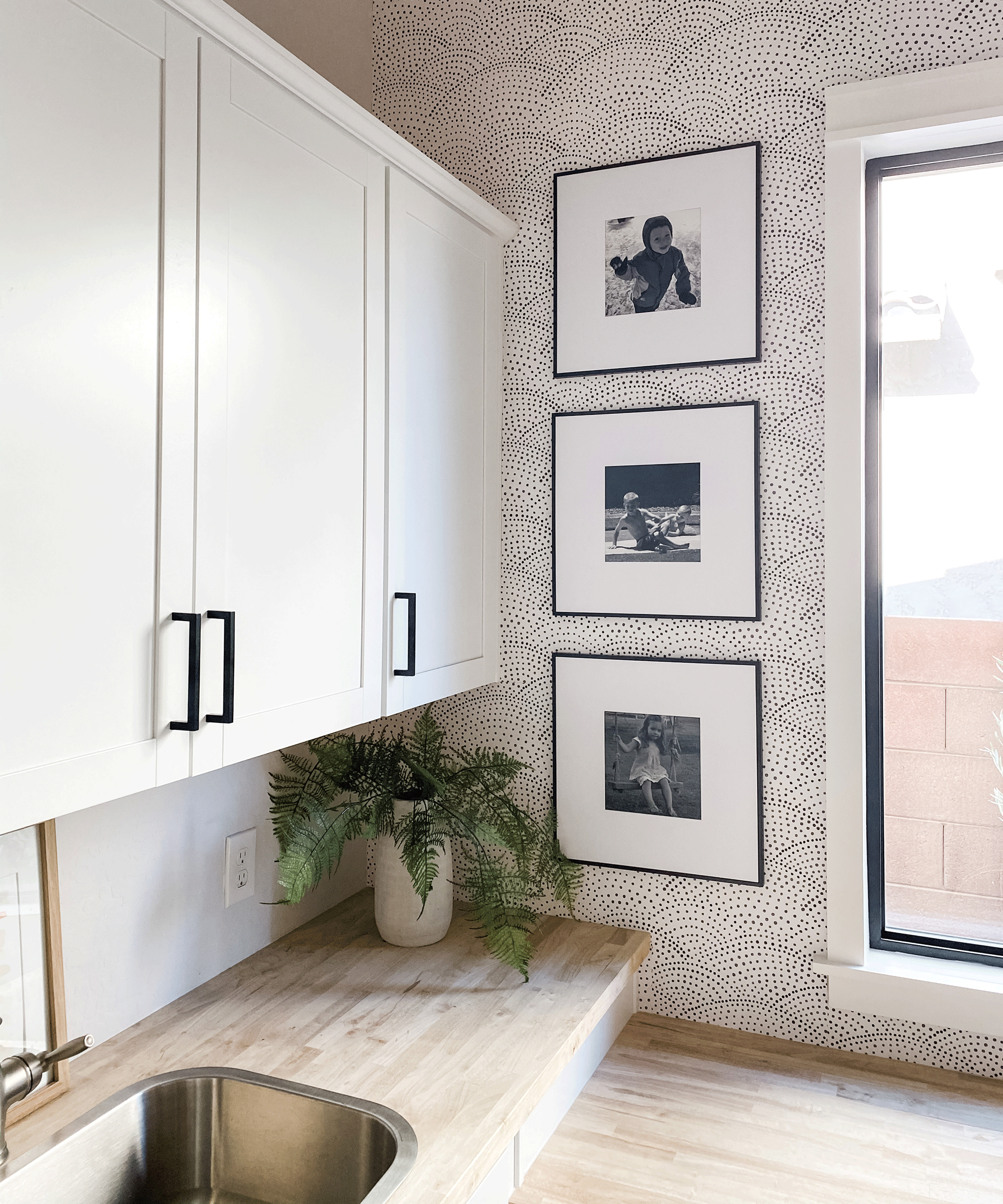 A kitchen with white cupboard decor, metal sink, wooden-effect worktops, trio of black and white framed wall art and monochrome spotted wallpaper with houseplant in corner