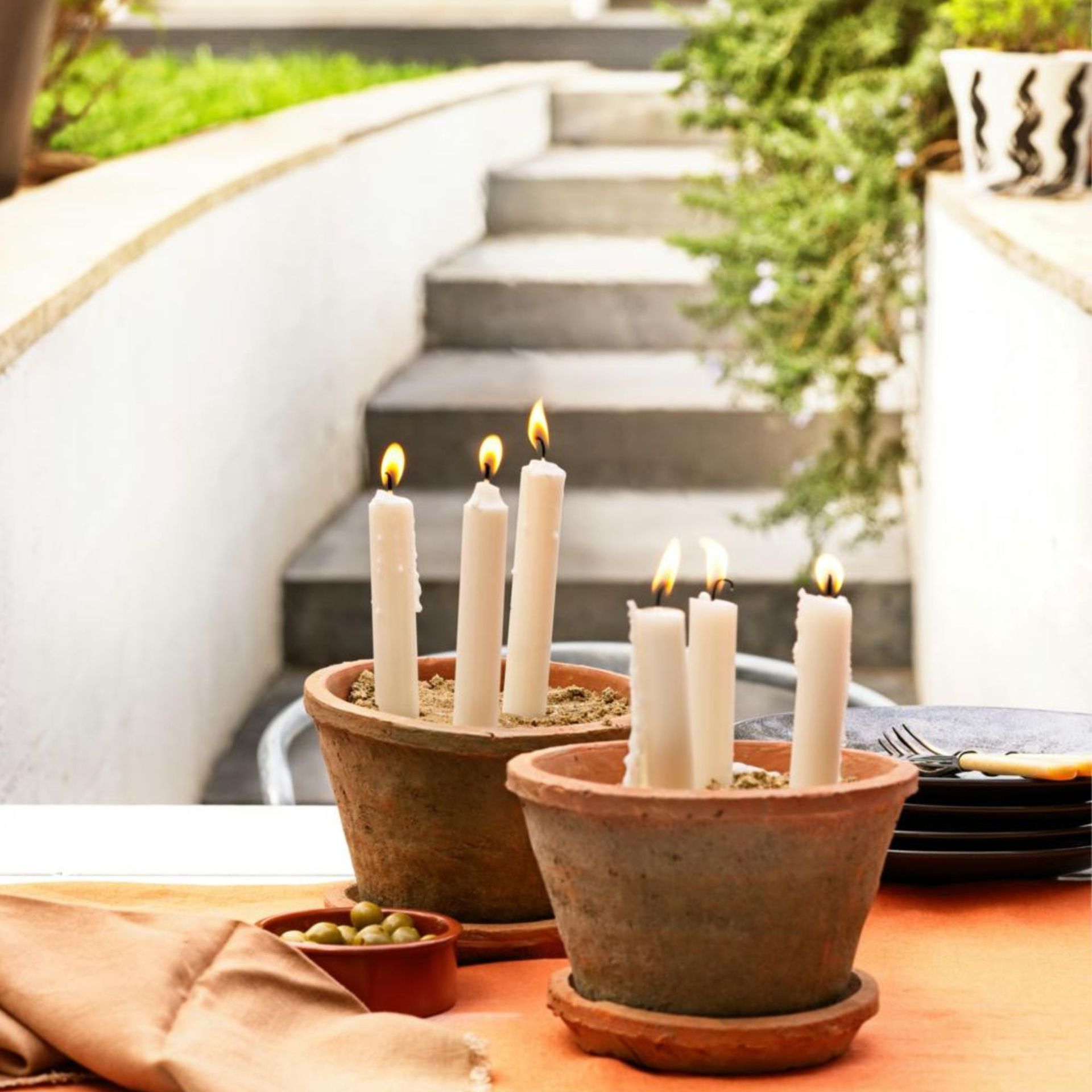 Candle planters using terracotta pots to make a simple patio display, a table with crockery and cutlery outdoors, with lit candles in old terracotta pots.