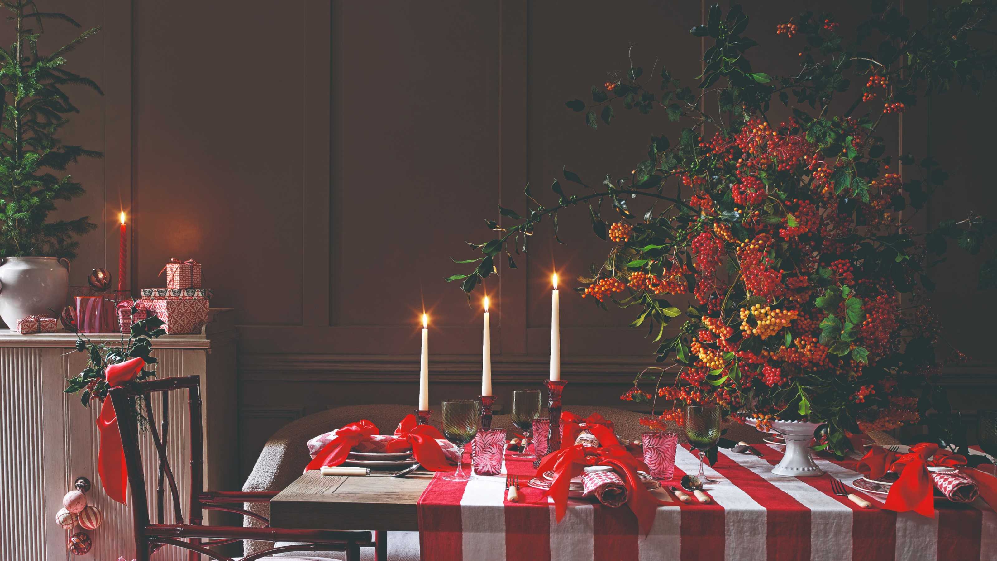 A dining table set and decorated for Christmas with a large centrepiece, dinner candles and red bows