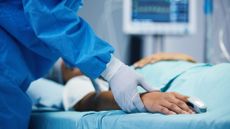 Close-up of a surgeon touching the hand of a patient who is lying in a hospital bed. Hospital machinery can be seen blurred in the background