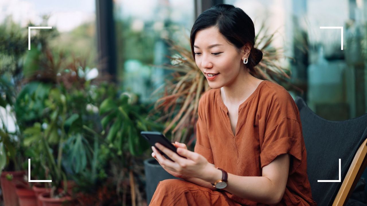 Woman sitting out on balcony surrounded by plants looking at the best journaling apps on her phone