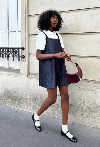 A woman's jean dress outfit with square-neck dress layered over white polo styled with white socks and black Mary Jane flats.