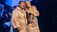 Guillermo and Witney Carson in brown trench coats ahead of their dance performance on Dancing With The Stars Week 4. They're under a blue umbrella and he has his arm around her. 