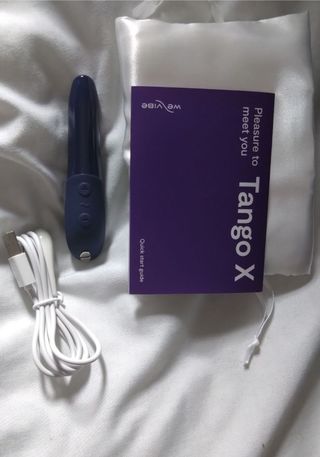 We-Vibe Tango X review image by Ness Cooper, we-vibe tango x in blue with satin storage bag, charging cable, and instruction booklet on white bedding.