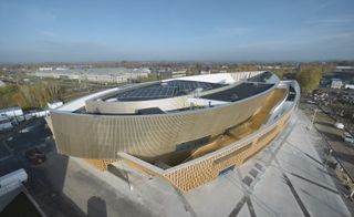 Day time aerial image, showing the curved design and structure of the golden brown anodized aluminium and tactile slats of Robinia wood framed building, with surrounding roads, cars, trees and street lights