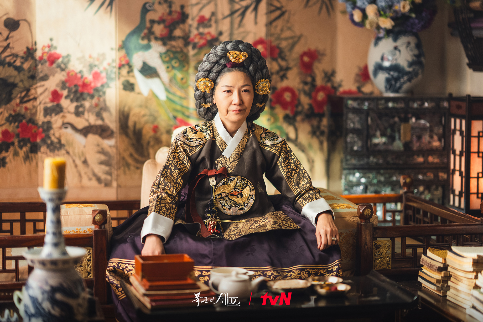 An older woman (Seo Yi-sook) wears a Joseon-era noblewoman outfit while sitting in a room at a dinner table, in &#039;Bon Appetit, Your Majesty.&#039;