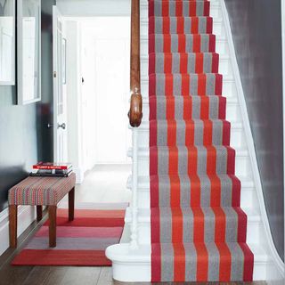 Red, grey and orange stair case runner on white stair case runner on white stair case next to hallway with bright stripe carpet on wooden floor