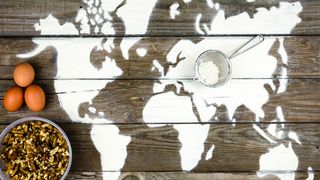 image of world map on a table. three eggs eggs sit to the left of north america. a bowl of grain is left of south america. on top of the border of europe and asia is a sieve with flower