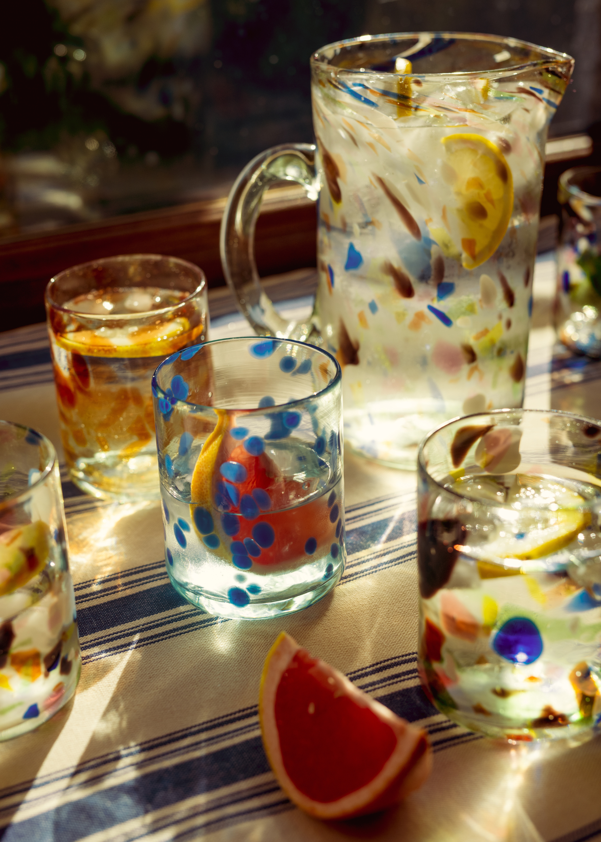 A striped dining table cloth holding glass-blown jugs and glasses alongside a slice of grapefruit