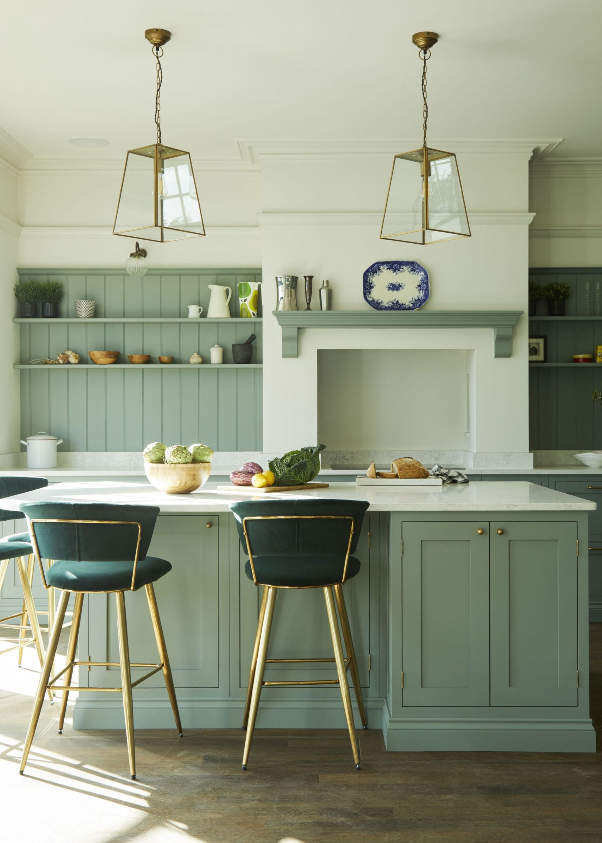 A sage green kitchen with an island and white countertops, two bar stools with gold legs and dark green seating with two gold-framed pendant lights hanging above