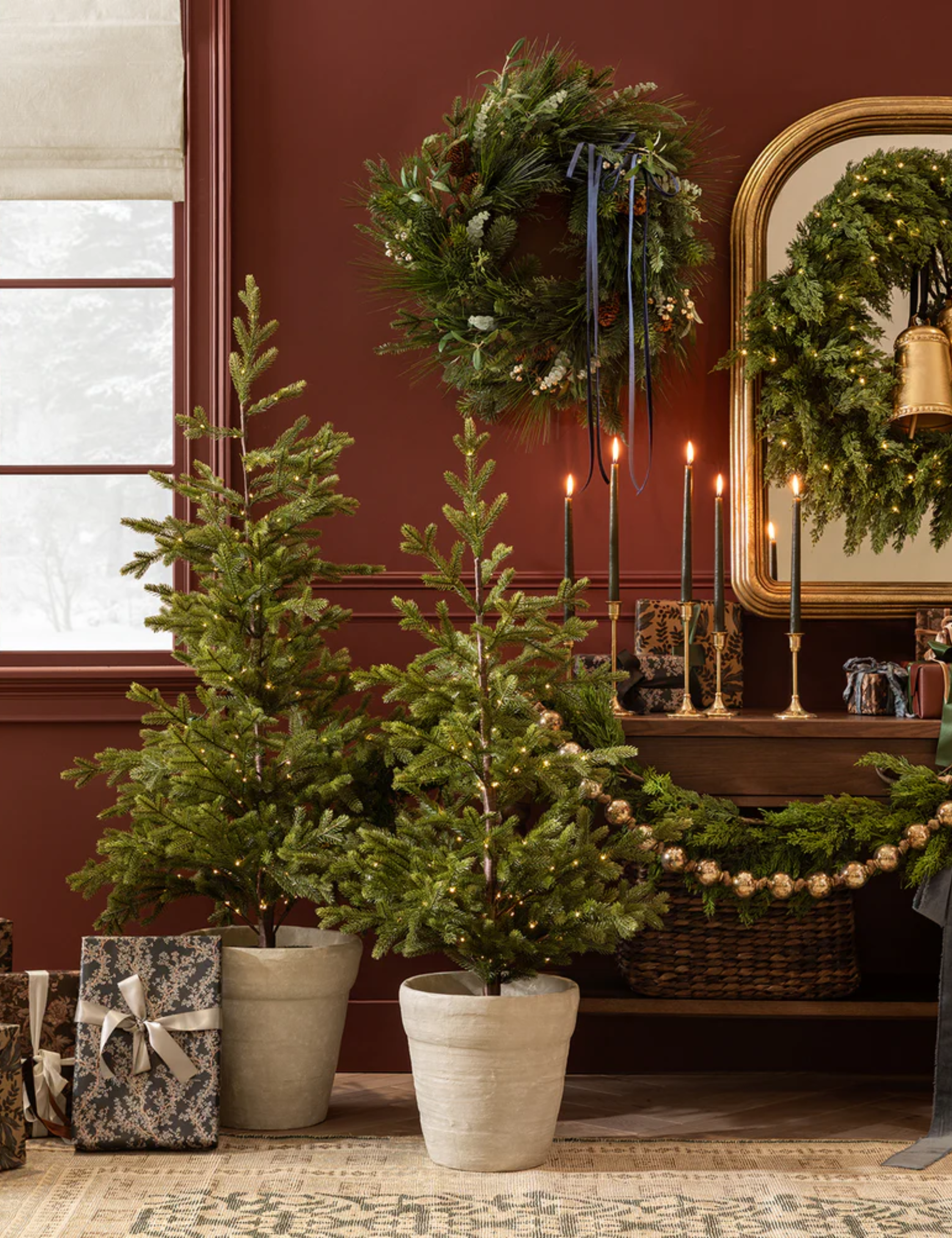 Shea McGee mini Christmas trees styled in a bordeaux living room area alongside Christmas wreaths, golden candlesticks, and wrapped presents.