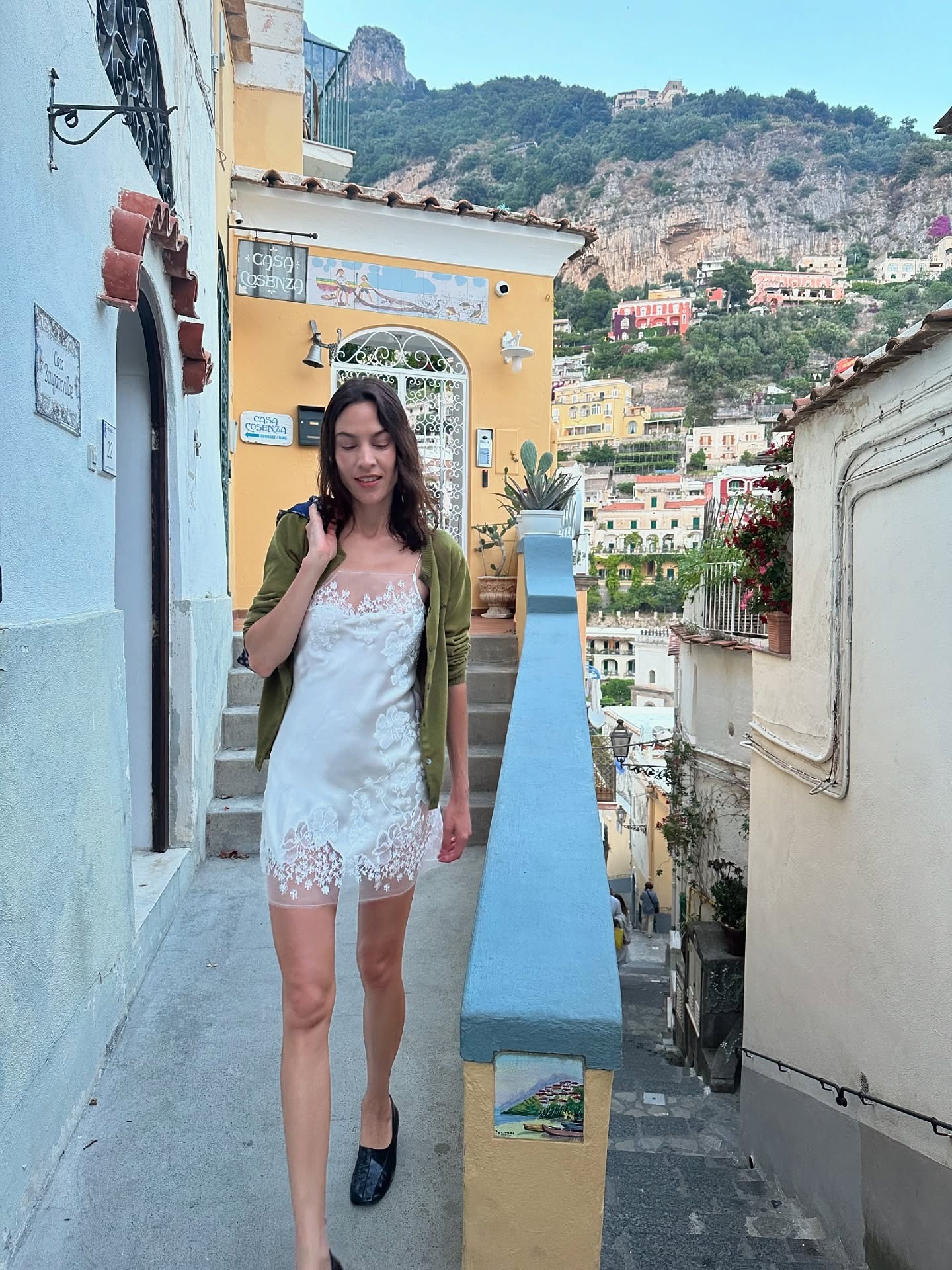 Alexa Chung walks down a pathway in Positano wearing a white satin lace slip dress with black square-toe flats and an olive green cardigan.