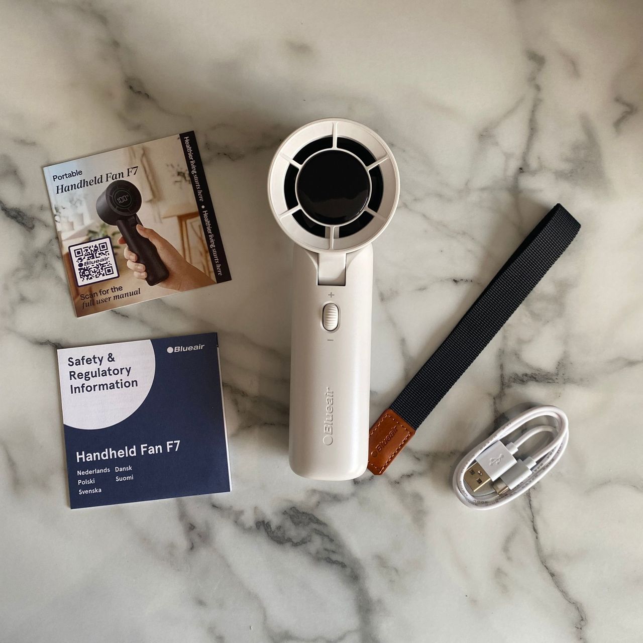 Flat lay of the Blueair AlwaysCool Portable Handheld Fan, instructional manuals and charging cable on a marble look worktop
