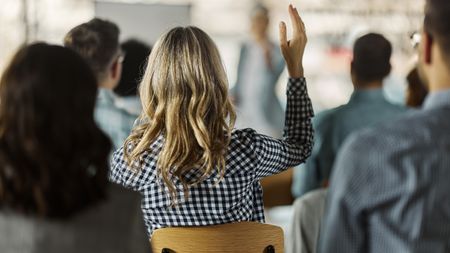 A worker raises her hand while in a meeting, her back to the camera.