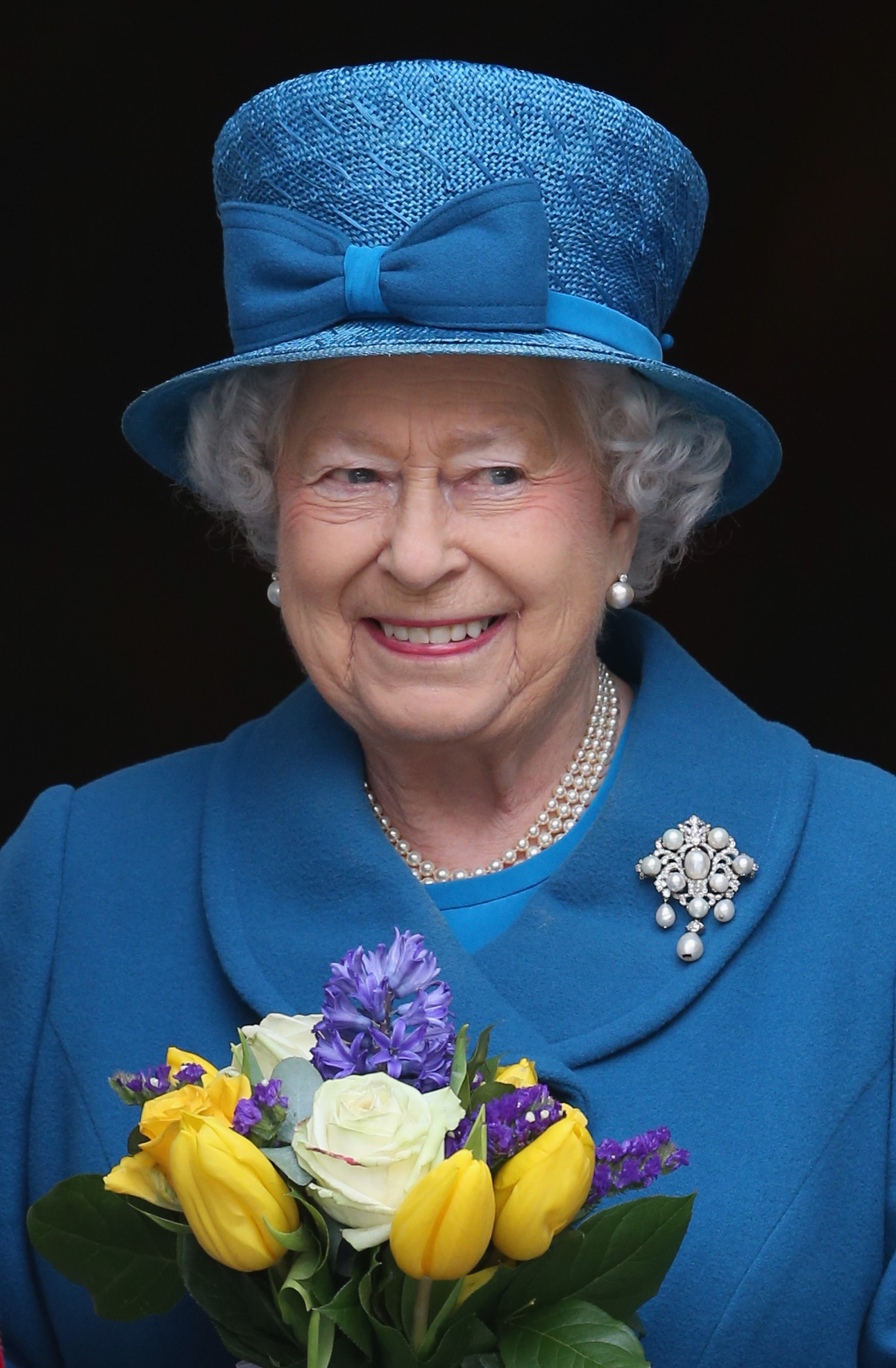 Queen Elizabeth wearing a blue cot and a hat holding flowers