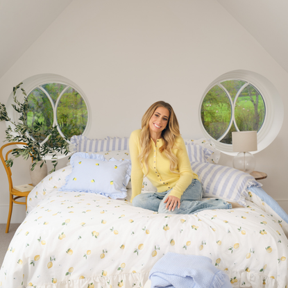 Stacey Solomon in blue jeans and yellow cardigan sat on double bed. The bedding is white, frilled, with yellow lemons printed on it. Behind are two circular windows looking out to a field and a plant.