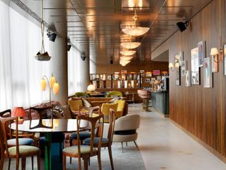 Alternative view of the lounge at White City House featuring grey flooring, wood covered walls, colourful wall art, curved pendant lights, wall lights and multiple lamps, round tables and seating in different colours. The bar area and restaurant can be seen in the distance
