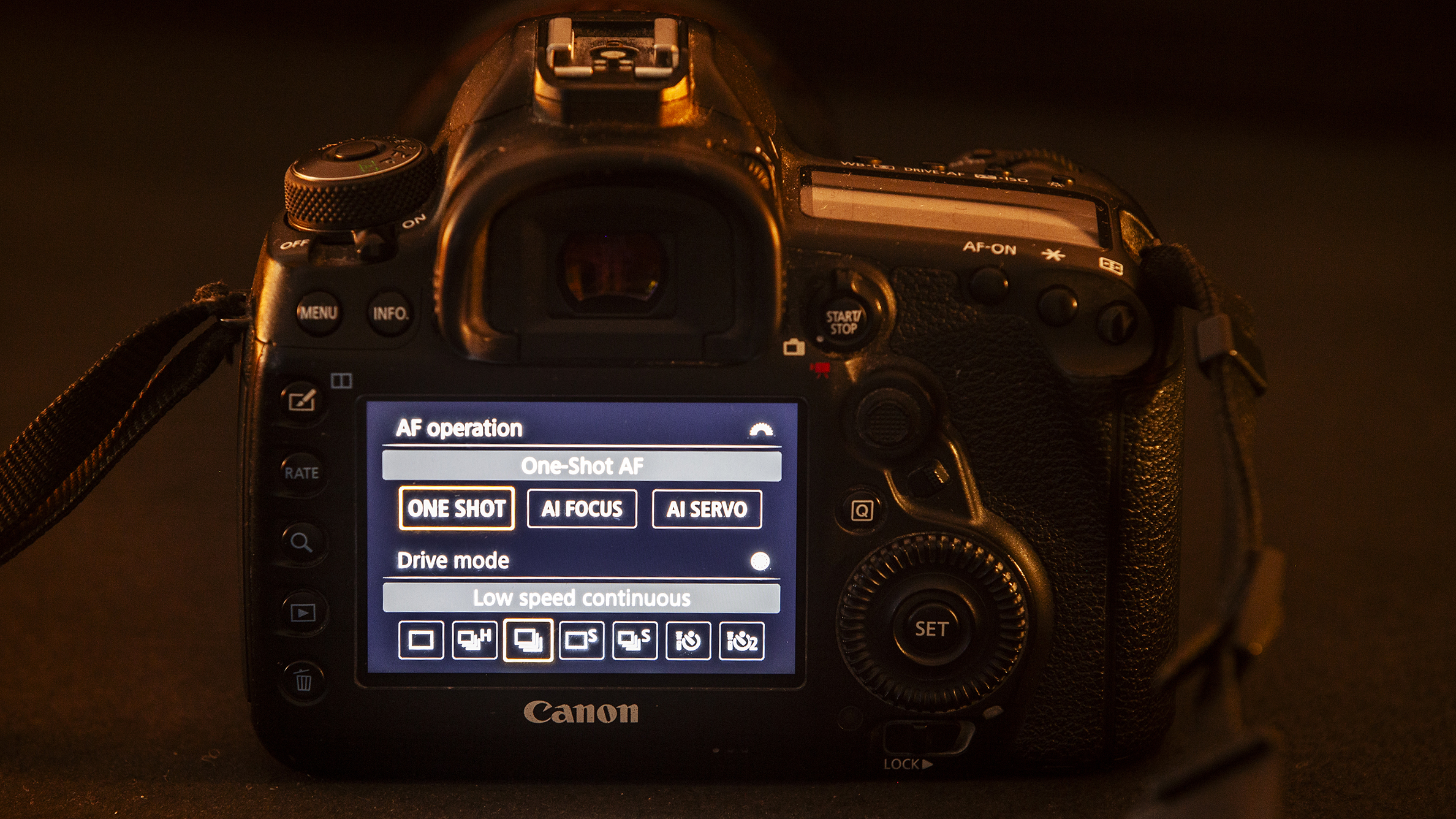 Rear view of a Canon DSLR camera in warm lighting. The screen displays AF operation settings with options like One Shot and AI Servo
