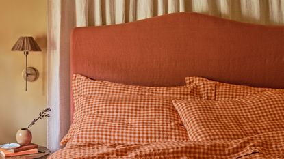 Close-up image of a bed with an orange cloth headboard and orange and peach gingham bedding. The walls are pale yellow, and there is a beige curtain behind the bed.