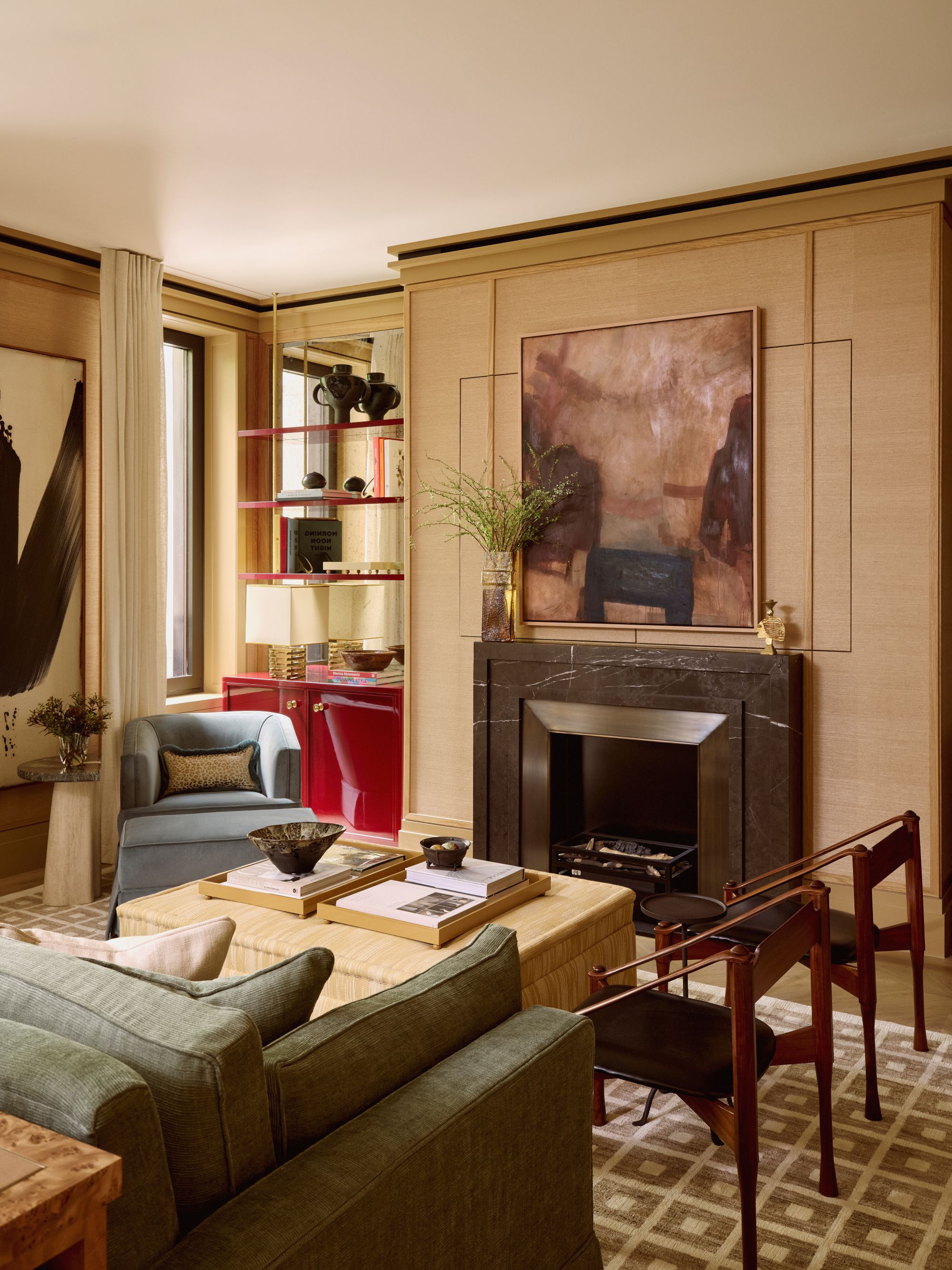 a sitting room with two wood framed chairs, a sage green couch, a tan ottoman, and a blue accent chair. Above the marble fireplace is an abstract painting. Adjacent is a red lacquer bookshelf 