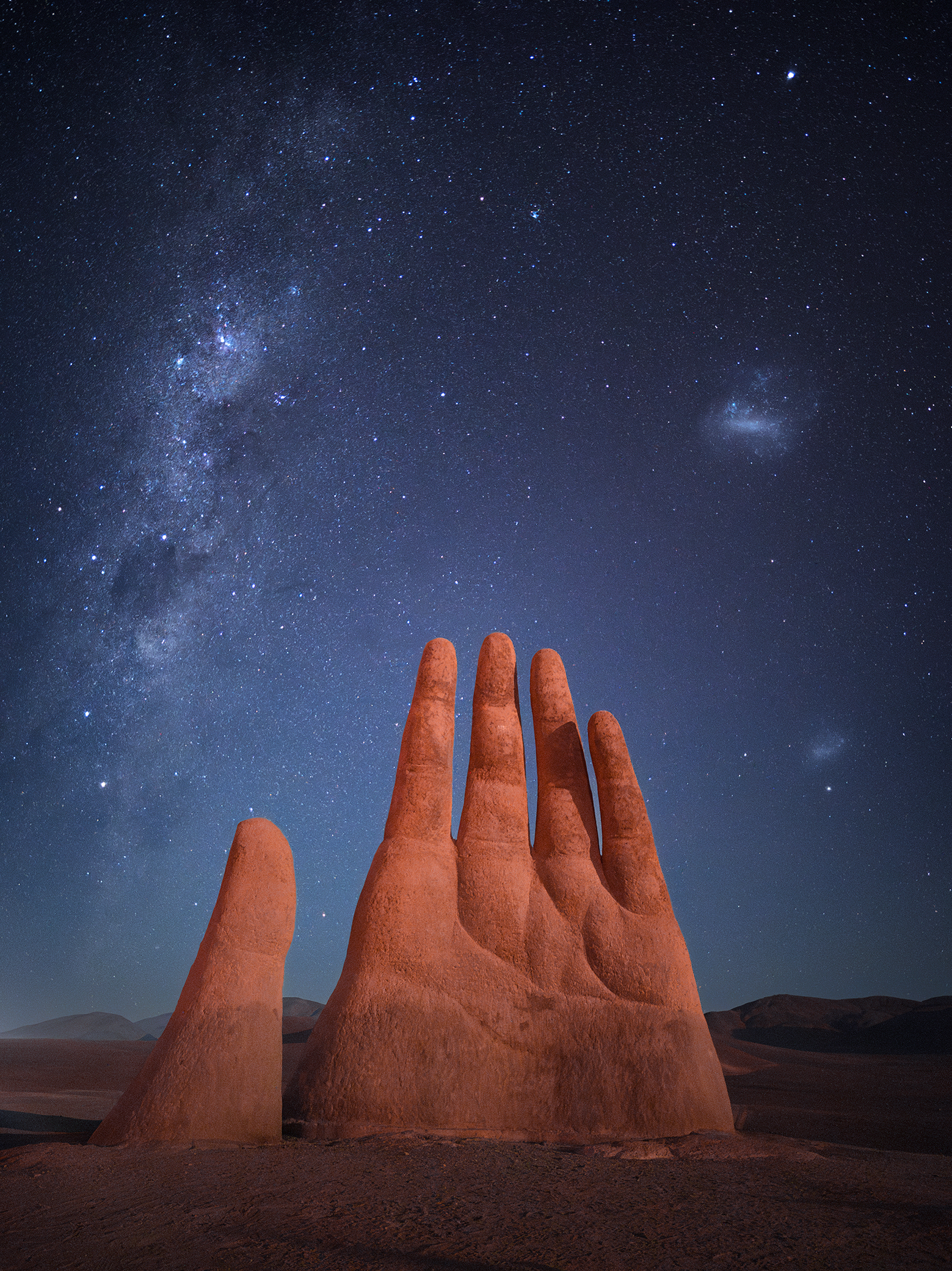 A large, surreal sculpture of a hand rises from the desert, under a starry night sky illuminated by the Milky Way