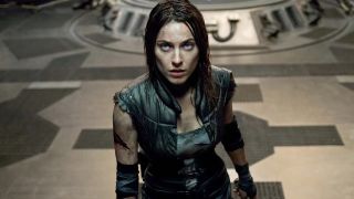 A still from the movie Pandorum. Here we see a woman with dark, shoulder length hair and blue eyes looking up. She is dressed in a leather vest, dirty white t-shirt, black elbow pads, and dark trousers. She is covered in dirt and has a wound on her right arm.