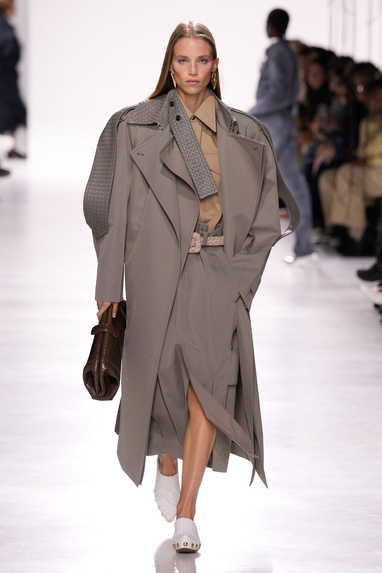 Bottega Veneta model walking in the S/S 26 show wearing a gray trench coat and carrying an east-west clutch bag.