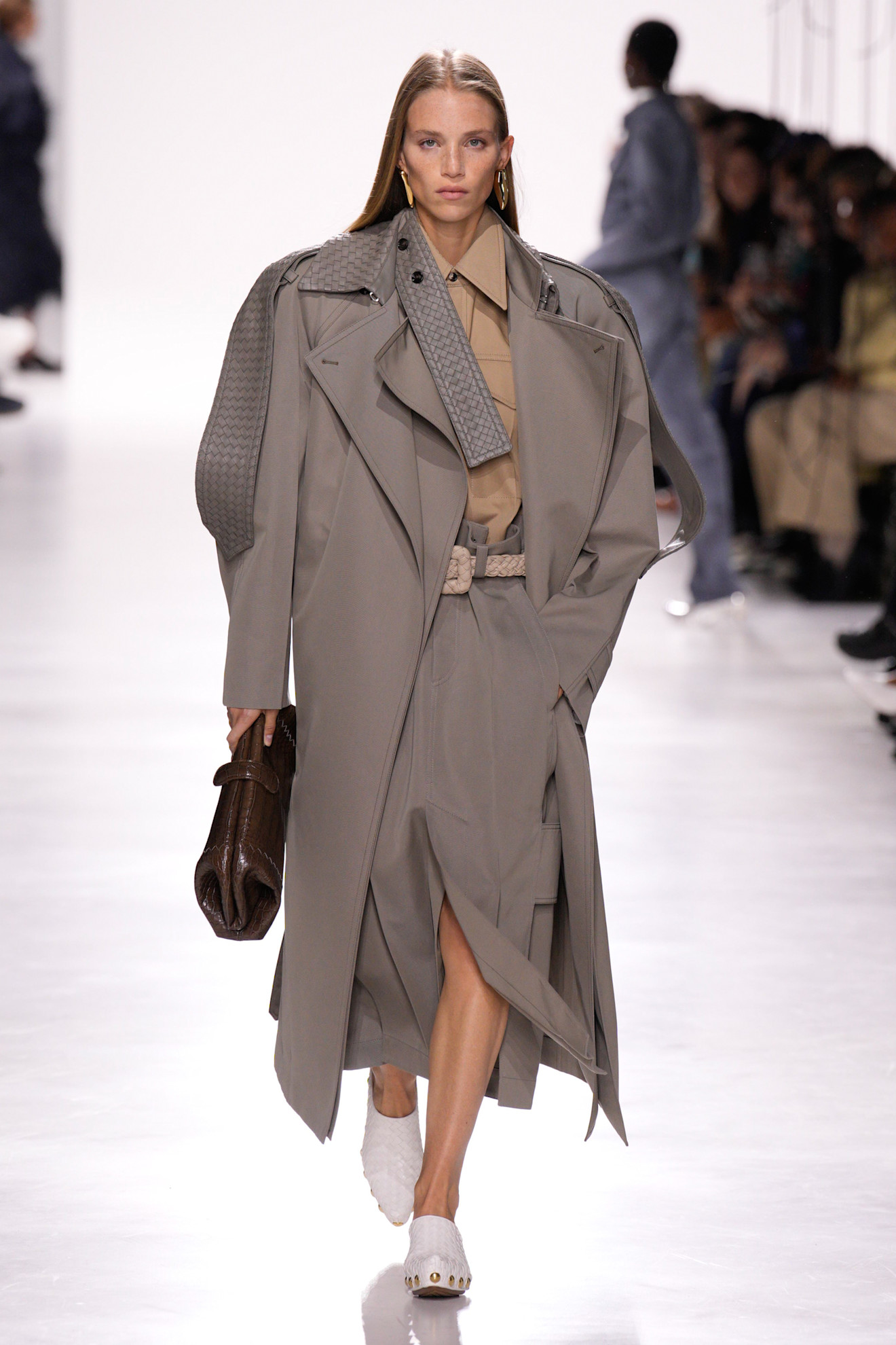 Bottega Veneta model walking in the S/S 26 show wearing a gray trench coat and carrying an east-west clutch bag.