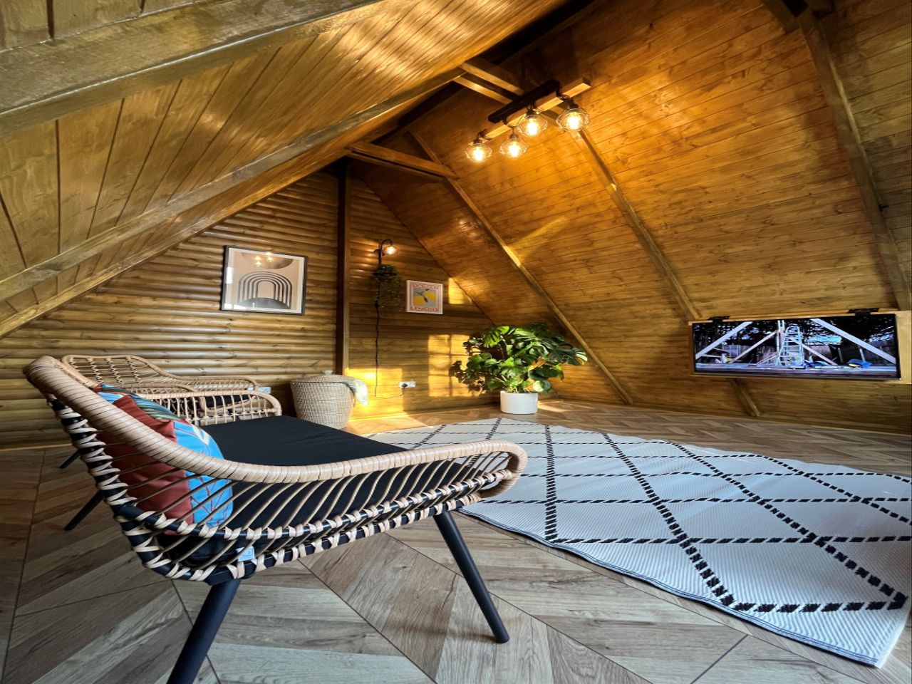 The interior of the chalet shows lighting fixtures above with a woven sofa chair and tv installed into the wall as well as small carpet covering a section of the wooden flooring