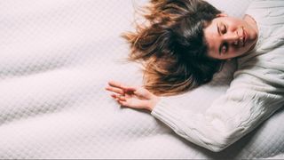 Aerial shot of a woman wearing a white jumper laying on a white mattress with her arm stretched over head and brown hair spread out.