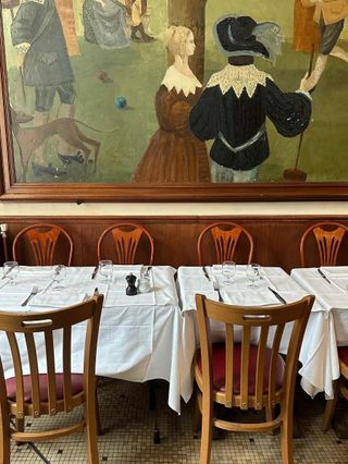 A classically decorated restaurant features a wall-large painting of a medieval public gathering, white table clothes, and wooden tables.