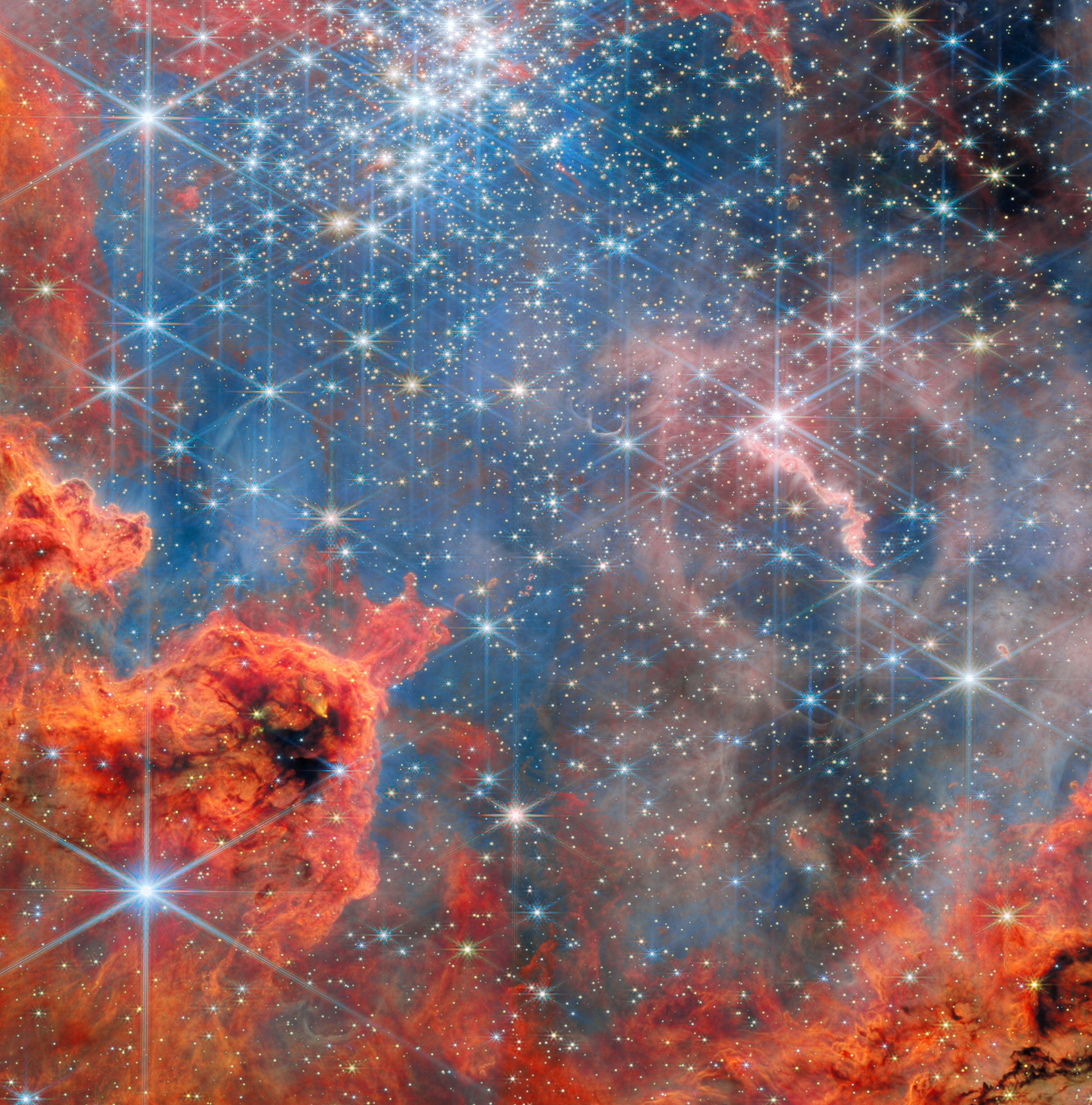 A cluster of stars inside a large nebula. The clouds of gas and dust are predominantly bright red in colour and wispy, akin to flames. They are clumped in the bottom-left corner. Other clouds, deeper in the cluster behind many of the stars, appear pale pink. The stars are concentrated in the top half of the image and are mostly small, bright white and six-pointed. They cast blue light over the nebula. Other stars with very long spikes surrounding them lie in the foreground.