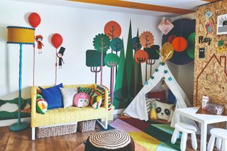 Spillett house: playroom with colourful tree wall stencil, hanging paper decorations, yellow sofa, white teepee, multicolour geometric rug