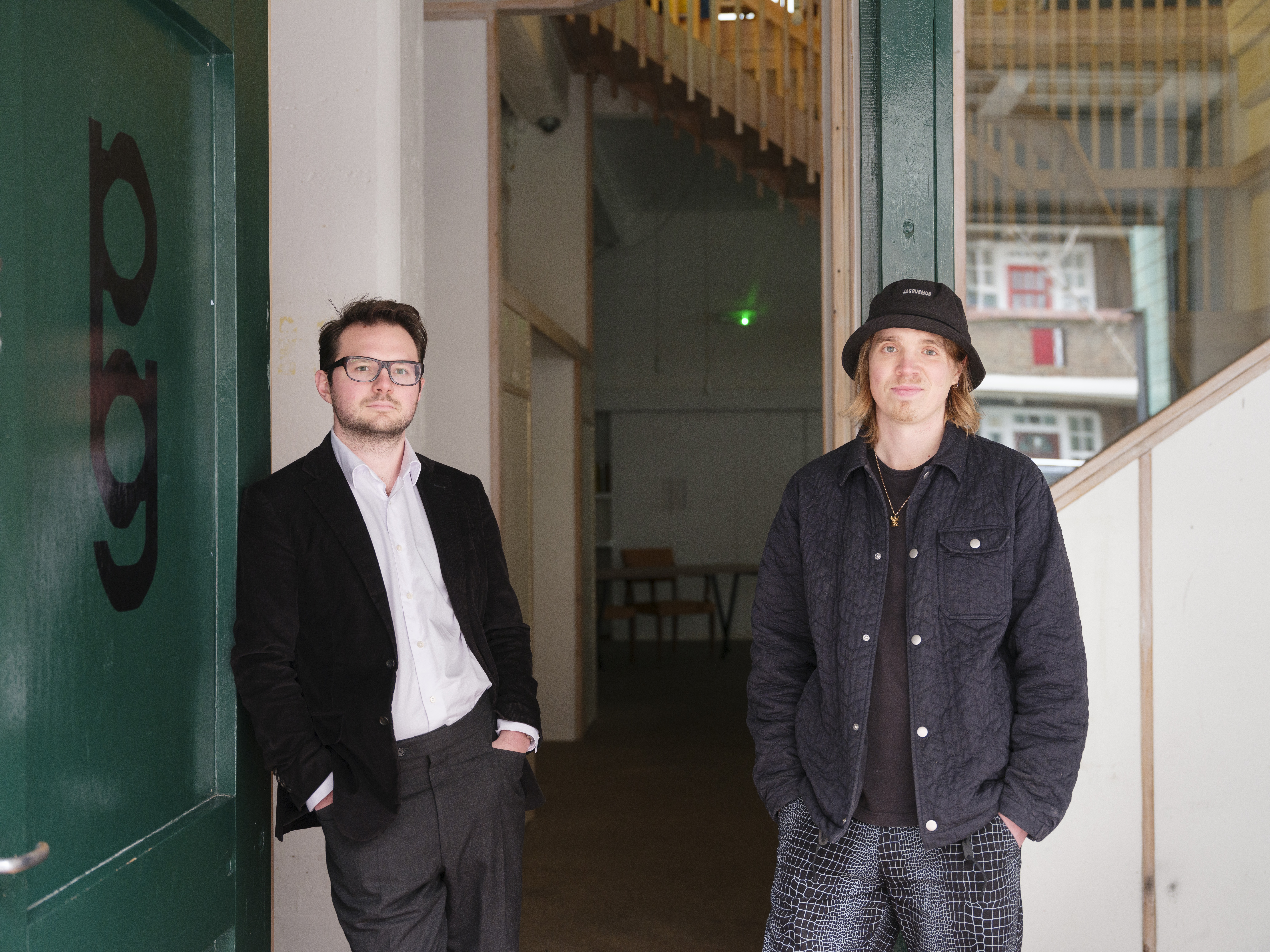 Two young men, one dressed in more elegant clothing, with a white shirt, black blazer, and gray suit trousers, the other wearing more casual attire in tones of black, gray, and brown, and a rain hat stand in front of the entrance of an industrial building housing a gallery.