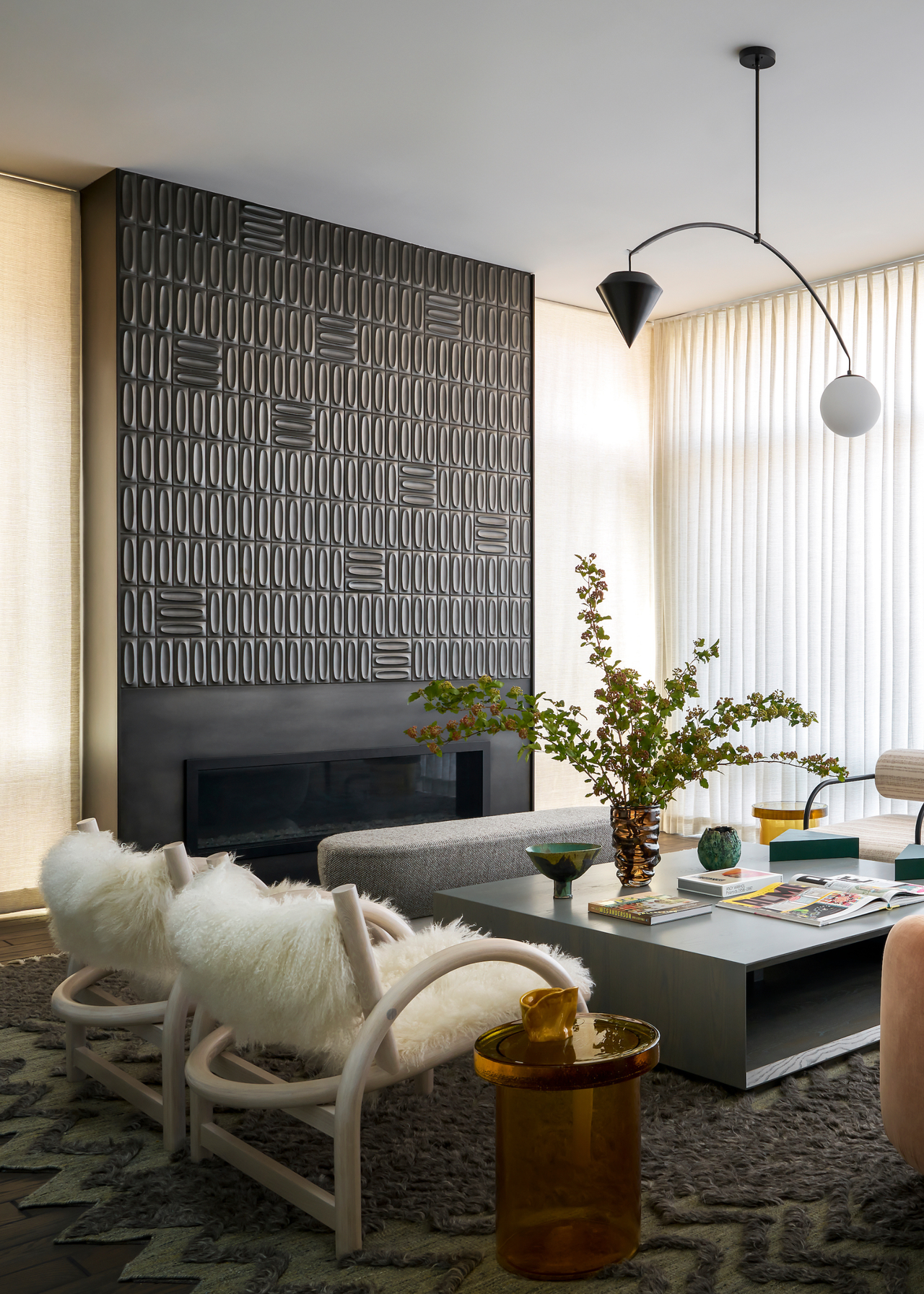 A living room with a black tile accent wall over a fireplace, a gray bench, a pair of white armchairs with shaggy seating, an amber glass side table