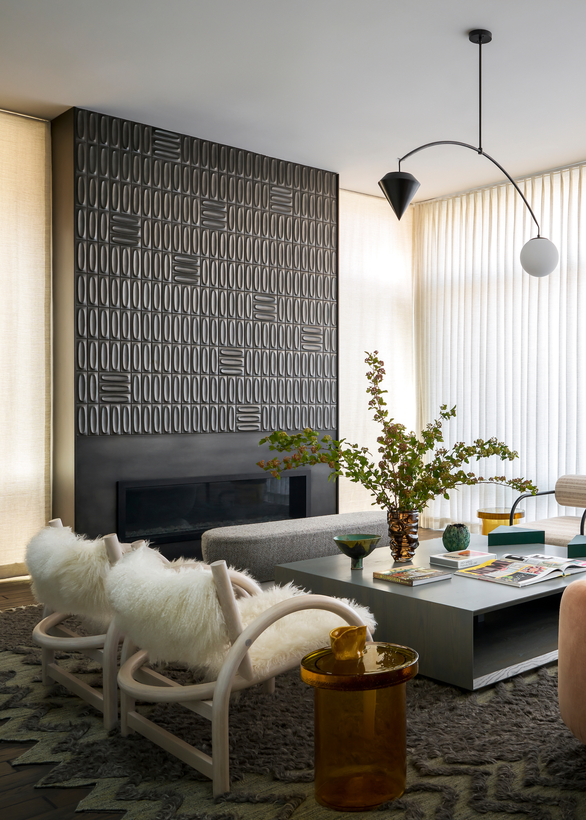 A living room with a black tile accent wall over a fireplace, a gray bench, a pair of white armchairs with shaggy seating, an amber glass side table