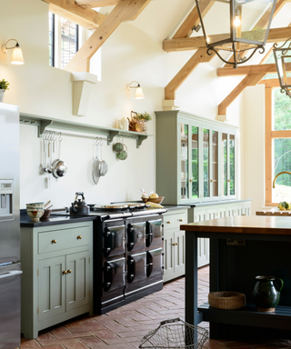 Black enamel Aga in a bright, country-style kitchen with wooden beams on pitch roof, sage green cupboards, an island and chevron stone floors.