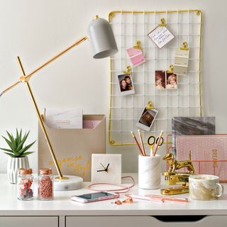 White desk next to a white wall with an angled gold desk lamp and a white metal grid memo board on the wall