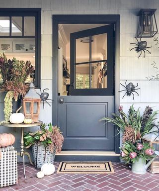 Halloween outdoor decor ideas with black dutch door, lantern style lighting, faux spider decor, flowers, pumpkins and doormat with herringbone floor design