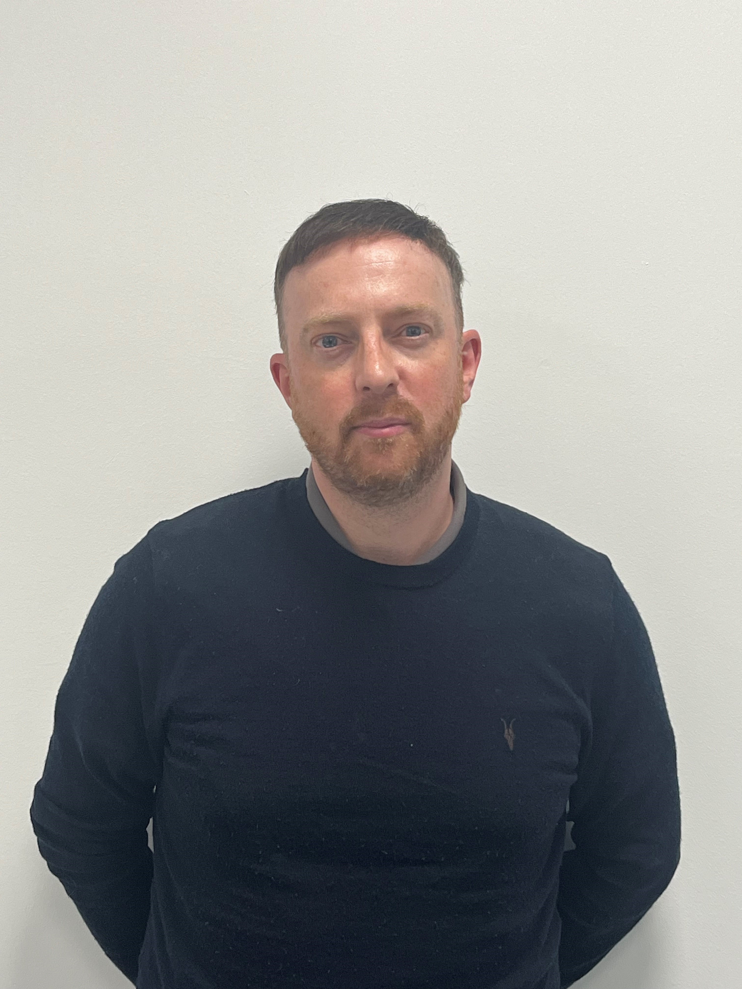 A man with a dark coloured jumper, brown hair and a brown beard standing in front of a white wall