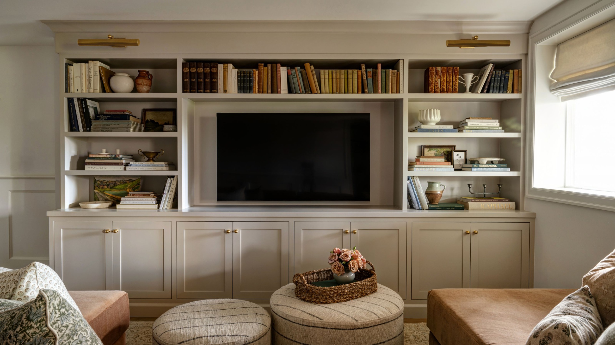 A basement living room with a wall of built-in shelving, including cupboards and open shelves, and a space for a mounted TV. Lots of books on display, brass picture lights, and a caramel-colored sofa with patterned cushions.
