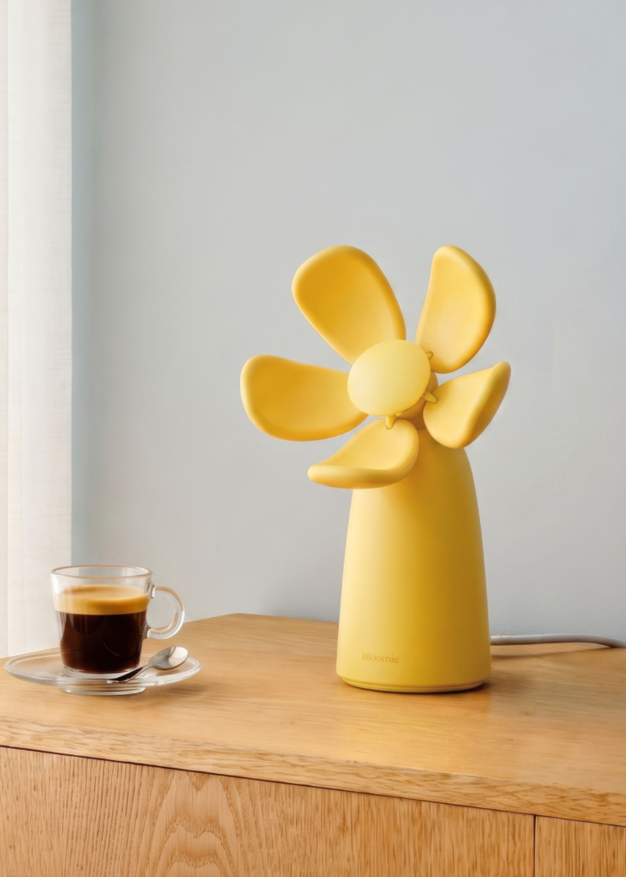A bright yellow flower-shaped air quality checker from Bloomie on a wood sideboard near an espresso in a clear cup and saucer