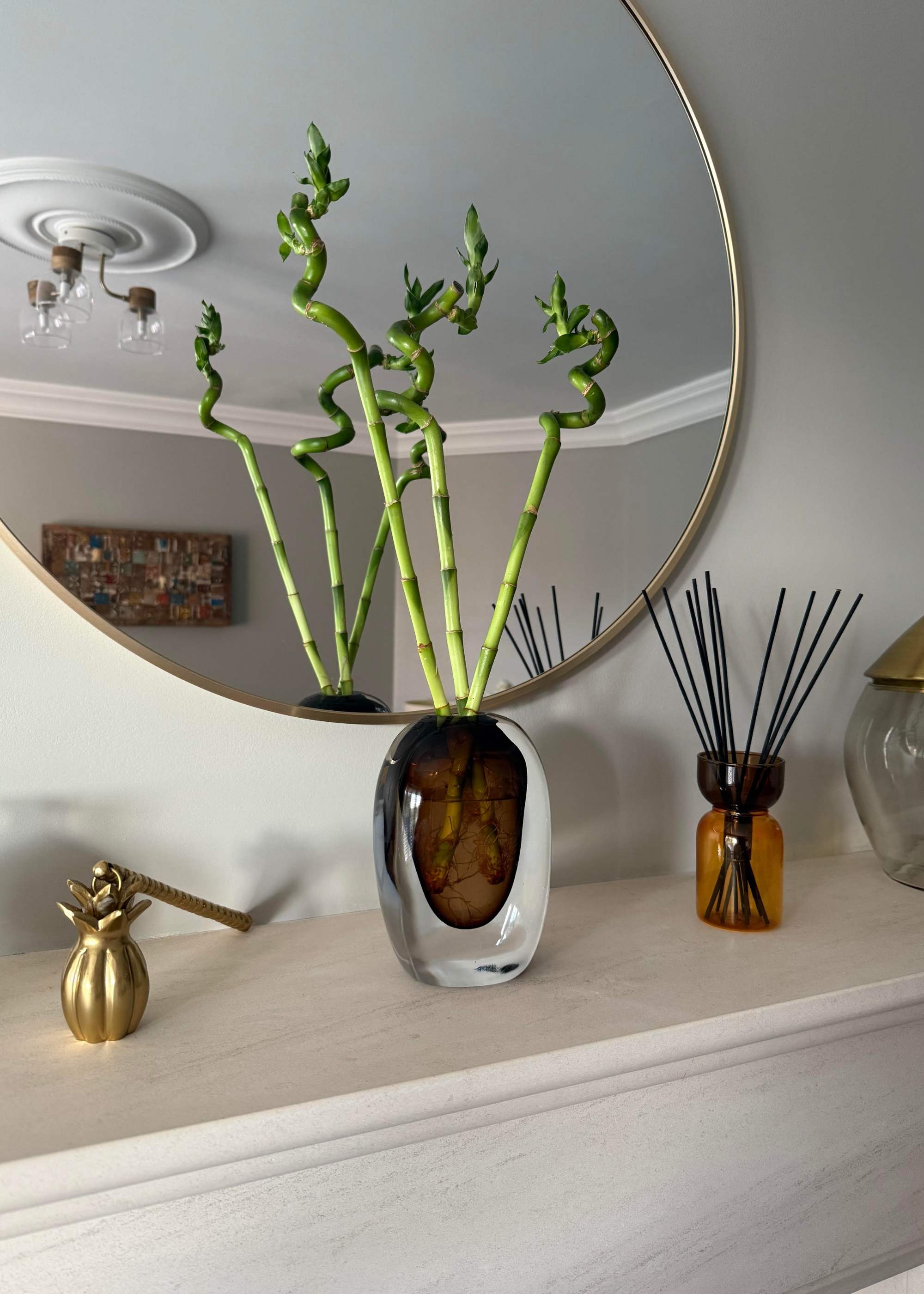 lucky bamboo spirals styled in a vase with water on a mantelpiece in front of a round mirror with a gold rim with a decorative candle holder on one side and a modern reed diffuser on the other