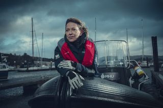 Nicola Walker in character as Annika, standing in a powerboat in the harbour wearing a navy windbreaker and a red lifejacket