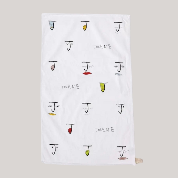 A white tea towel with stylized faces in black with yellow, red, and light blue, pink, and red mouths sits against a pale grey background.