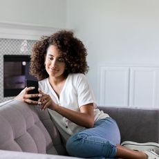 Woman smiles as she uses her phone while sat on a sofa.